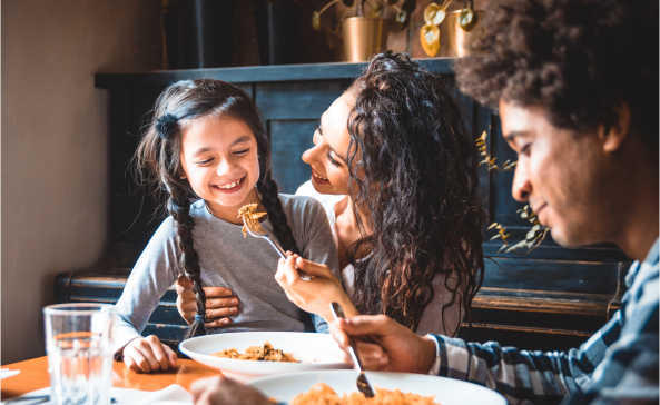 A young family sitting around the dinner table while the mother tries to feed her her daughter who is sitting on her lap.