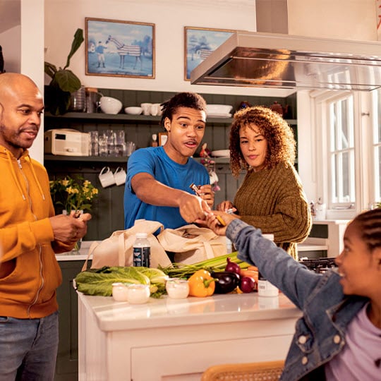 A photo of a family in a kitchen interacting around the kitchen island. Kitchen island includes fresh produce from a recent shopping trip.