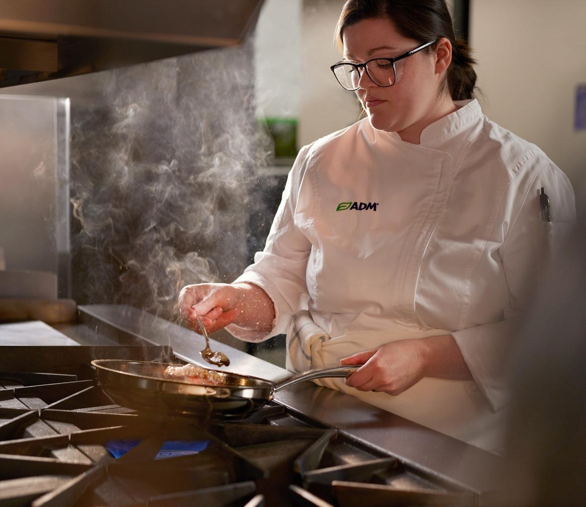 Female chef wearing white ADM coat cooking with frying pan on stove in culinary kitchen